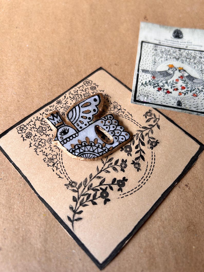 May include: A decorative bird-shaped button with a crown, featuring white, gold, and black details, rests on a diamond-shaped card with floral and leaf patterns. A small card with two birds and floral details is also present.