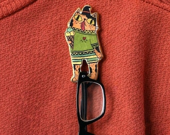 May include: A whimsical cat-shaped pin with a green sweater and ice cream cone design, paired with black-framed eyeglasses. The pin is attached to a rust-colored sweater, creating a playful contrast. The cat is wearing a hat and has its tongue out.