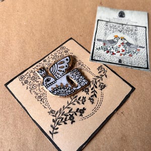 May include: A decorative bird-shaped wooden pin with a crown, black and white details, and gold accents, set on a square card with floral and leaf designs. A second card with bird illustrations is also present.