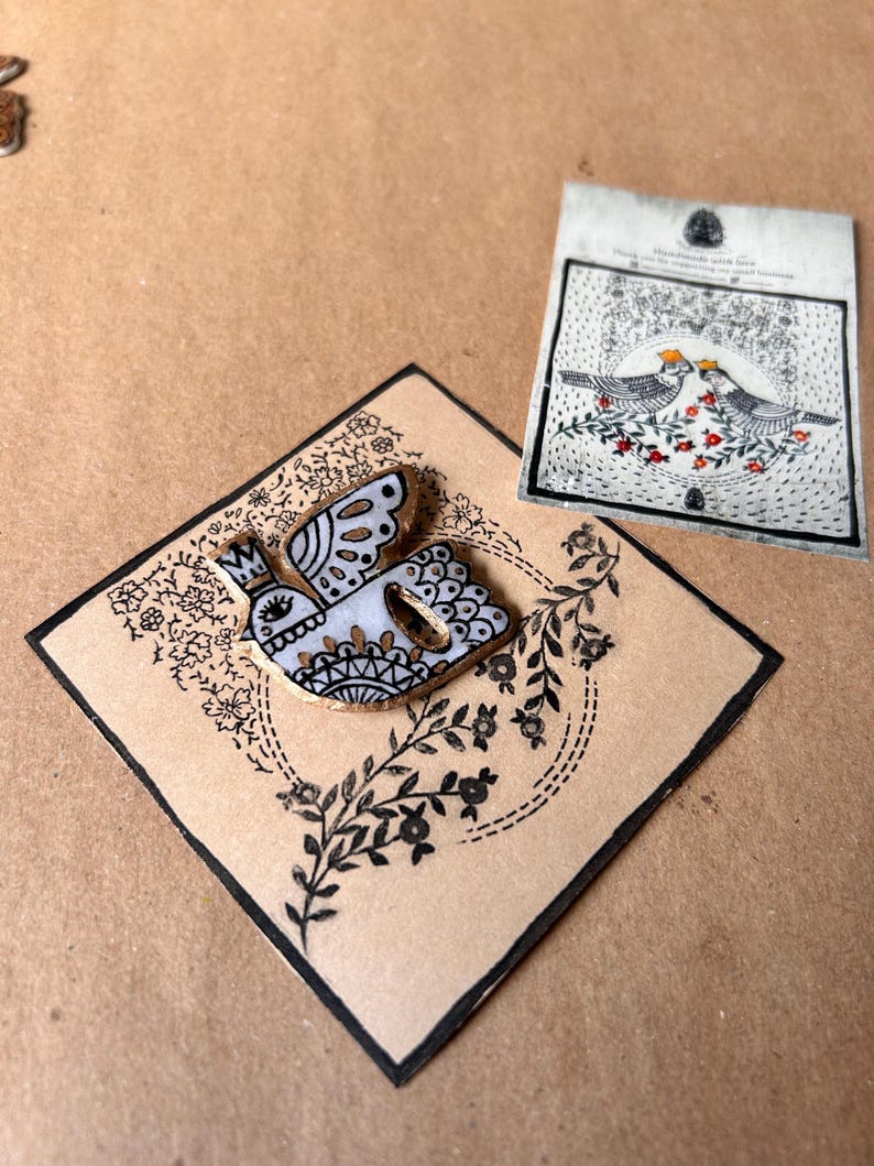 May include: A decorative bird-shaped wooden pin with a crown, black and white details, and gold accents, set on a square card with floral and leaf designs. A second card with bird illustrations is also present.