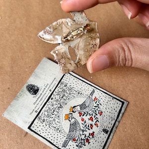 May include: A handmade, abstract-shaped brooch with a gold pin, held by a hand. The brooch is beige with a textured surface. A card with a black and white bird illustration and text is below the brooch.