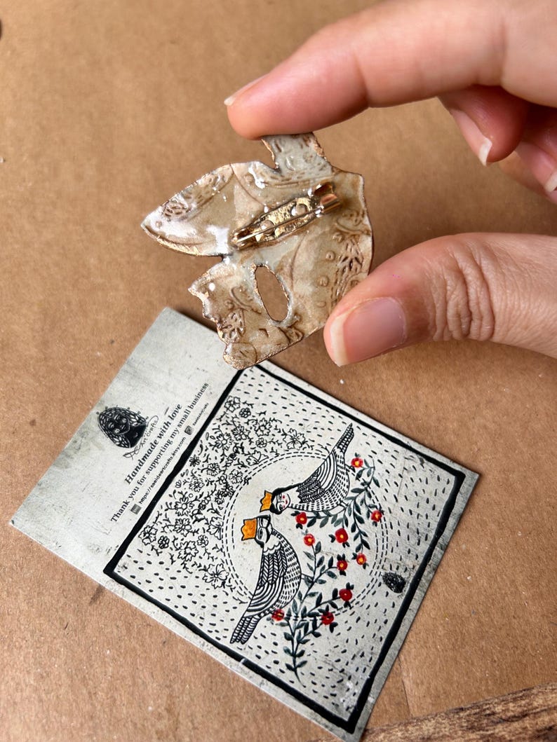 May include: A handmade, abstract-shaped brooch with a gold pin, held by a hand. The brooch is beige with a textured surface. A card with a black and white bird illustration and text is below the brooch.