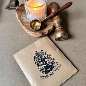 May include: A lit candle in a glass jar, a small brass ladle, and a wooden handled wax stamp rest on a wooden tray. A small brown paper package with a black illustration and the words "Art Crafts" is in the foreground.