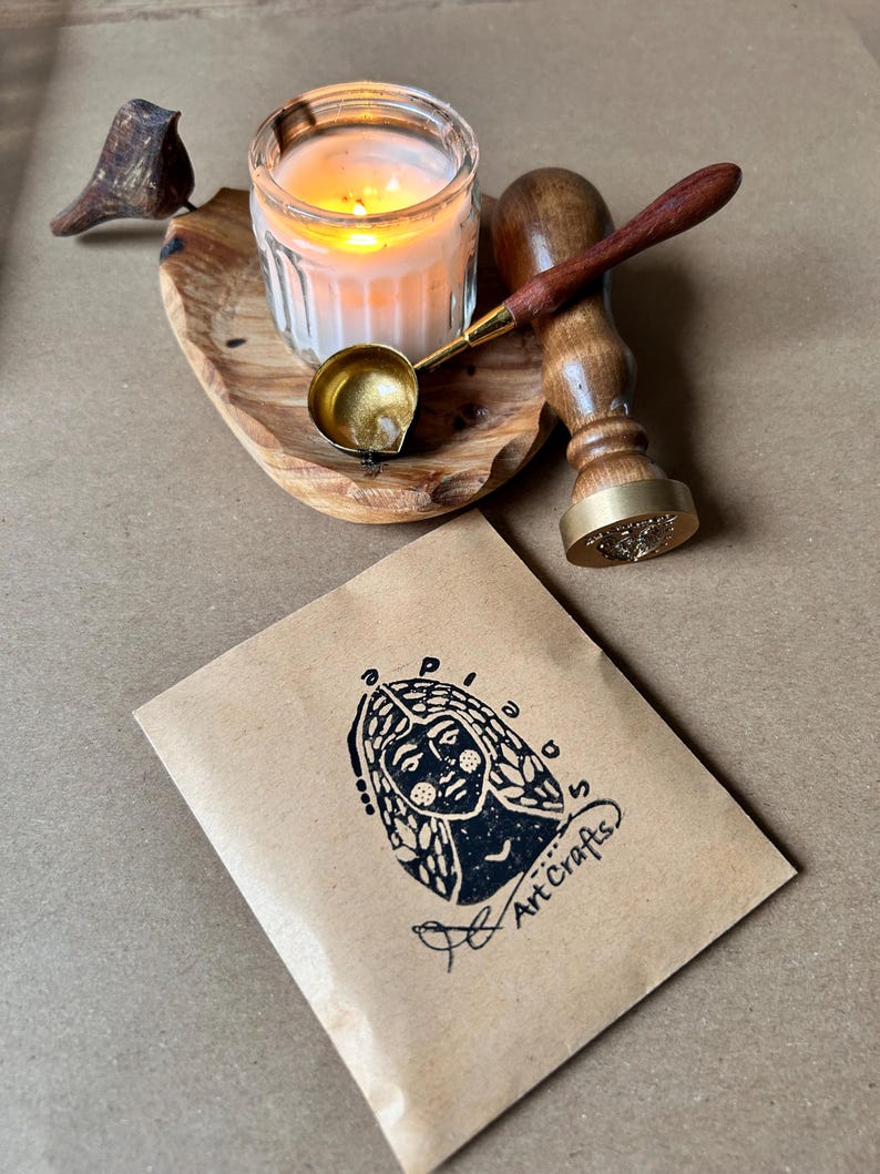 May include: A lit candle in a glass jar, a small brass ladle, and a wooden handled wax stamp rest on a wooden tray. A small brown paper package with a black illustration and the words "Art Crafts" is in the foreground.