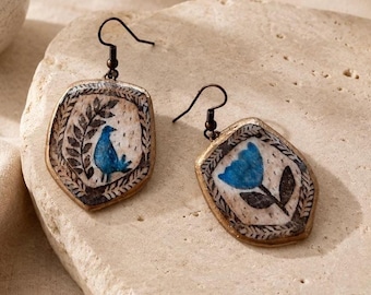 May include: A pair of dangle earrings featuring irregular, shield-shaped pendants with a textured, off-white surface. One earring displays a blue bird next to a leafy branch, while the other shows a blue flower. Both have dark metal hooks.