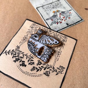 May include: A white and gold bird-shaped brooch with black decorative details, set on a beige card with floral designs. A matching card with two birds and floral accents is also visible. The brooch has a gold border.