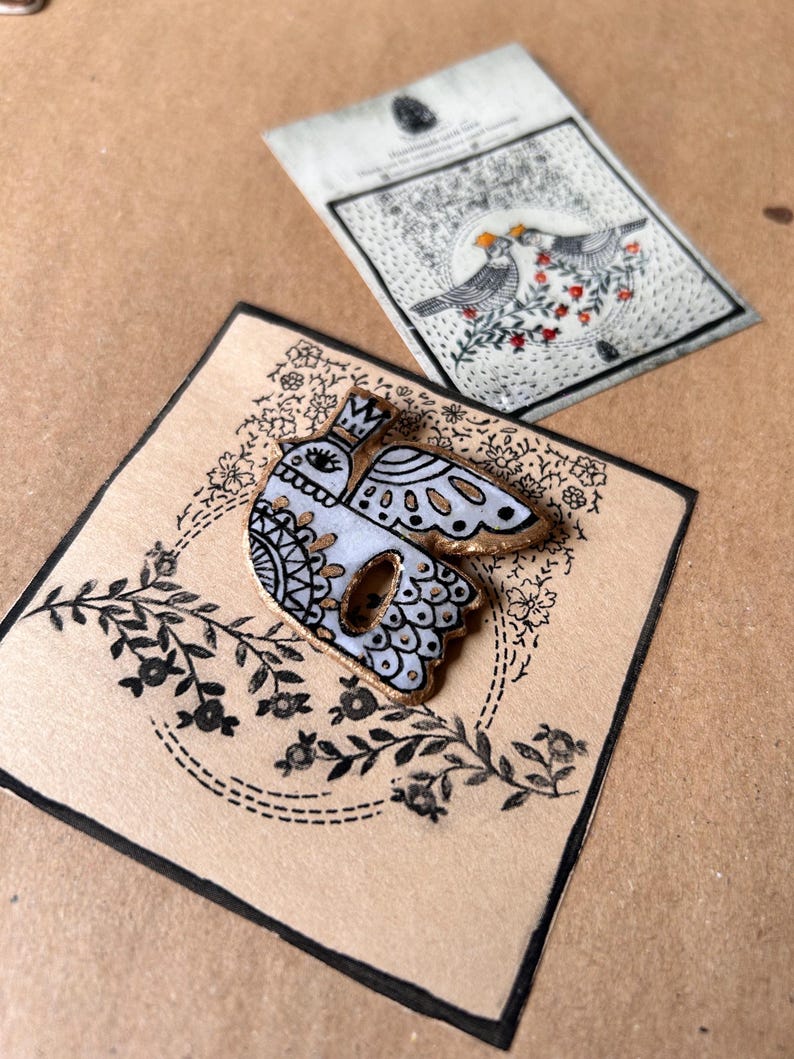 May include: A white and gold bird-shaped brooch with black decorative details, set on a beige card with floral designs. A matching card with two birds and floral accents is also visible. The brooch has a gold border.