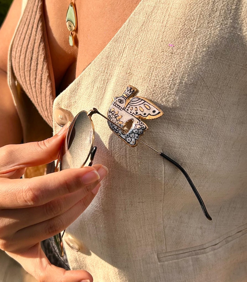 May include: A decorative bird-shaped pin with a white, gold, and black design, holding a pair of eyeglasses. The pin is attached to a beige vest. An eye-shaped pendant hangs from a necklace.