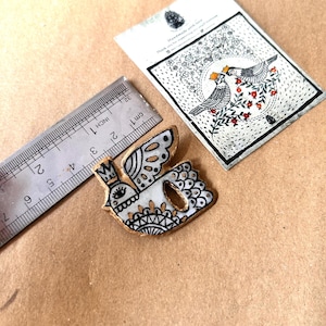 May include: A handmade, white and gold bird-shaped brooch with a crown, wings, and intricate black detailing. A ruler shows the brooch is approximately 2 inches long. A card with a bird illustration is also present.