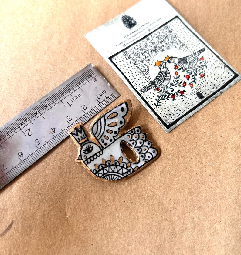 May include: A handmade, white and gold bird-shaped brooch with a crown, wings, and intricate black detailing. A ruler shows the brooch is approximately 2 inches long. A card with a bird illustration is also present.