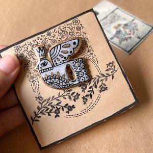 May include: A white and gold bird pin with a crown, black details, and a floral design on a tan card. The pin is on a card with a black border and floral accents. A second card with a bird illustration is in the background.