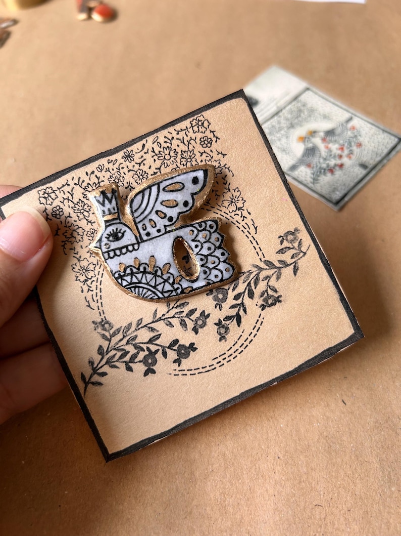 May include: A white and gold bird pin with a crown, black details, and a floral design on a tan card. The pin is on a card with a black border and floral accents. A second card with a bird illustration is in the background.