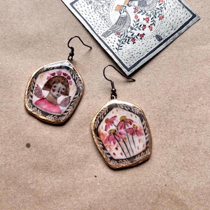 Può includere: Orecchini artigianali fatti a mano con disegni dipinti unici. Uno presenta una figura con ali e abito rosa, l'altro fiori rosa. Entrambi hanno un bordo dorato e ganci in metallo scuro.