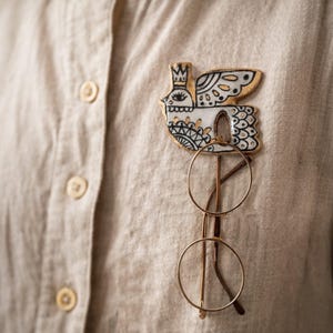May include: A decorative bird-shaped pin with gold accents, black details, and a crown, attached to a beige shirt. Below the pin, a pair of round, gold-rimmed glasses is displayed. The pin and glasses create a unique accessory combination.