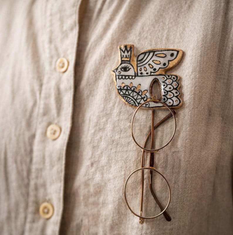 May include: A decorative bird-shaped pin with gold accents, black details, and a crown, attached to a beige shirt. Below the pin, a pair of round, gold-rimmed glasses is displayed. The pin and glasses create a unique accessory combination.