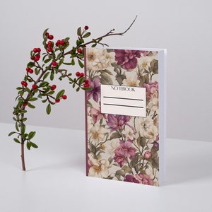 Könnte beinhalten: Ein Blumen-Notizbuch mit einem burgunderfarbenen Rücken und einem weißen Einband, auf dem das Wort "NOTEBOOK" gedruckt ist. Das Notizbuch liegt auf einer weißen Oberfläche mit einem Zweig mit roten Beeren und grünen Blättern dahinter.