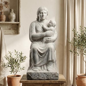 May include: A marble statue of a seated woman holding a baby. The sculpture is light gray and white, with detailed carving of the figures' clothing and features. The statue is placed on a wooden table, with potted plants in the foreground.