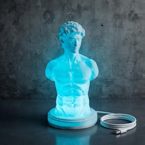 May include: A sculptural lamp shaped like a male bust, illuminated with a bright blue light. The figure features detailed facial features and a muscular torso, resting on a circular base with an attached white USB power cable extending to the side.