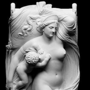 Puede incluir: Escultura blanca que representa a una mujer y un niño. La mujer, desnuda, tiene el pelo largo y un tocado. El niño se estira hacia ella. La escultura está colocada sobre un fondo de tela drapeada, sobre un fondo negro.
