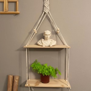 May include: A macrame hanging shelf with two wooden shelves. The top shelf holds a white bust sculpture, while the bottom shelf displays a potted green plant. The shelf is suspended by white rope and wooden beads.