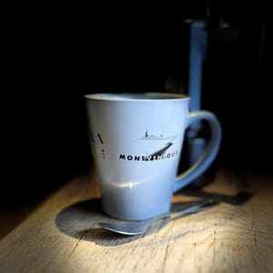 Pode incluir: Uma caneca de cerâmica branca com uma ilustração gráfica da paisagem do Mont Ventoux e o texto MONT VENTOUX impresso em preto e vermelho. A caneca tem uma asa curva e é acompanhada por uma pequena colher de metal de cor prateada.
