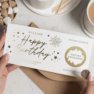 Może przedstawiać: Biała kartka urodzinowa ze złotymi literami i gwiazdkami. Na kartce widnieje napis "Happy Birthday" i imię "William". Pole zdrapki ujawnia prezent "Driving Experience". Moneta jest trzymana w pobliżu pola zdrapki.