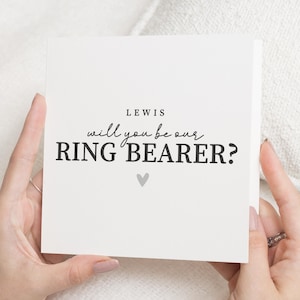 May include: A square, cream-coloured card held by hands, with the text "LEWIS will you be our RING BEARER?" in black. A small grey heart is centered below the text. The card is on a white background.