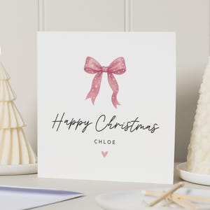 Könnte beinhalten: Eine weiße Weihnachtskarte mit einer rosa Schleifenillustration und dem Text "Happy Christmas" und dem Namen "Chloe". Zwei brennende, weiße, baumförmige Kerzen flankieren die Karte. Ein Teller mit Streichhölzern befindet sich im Vordergrund.