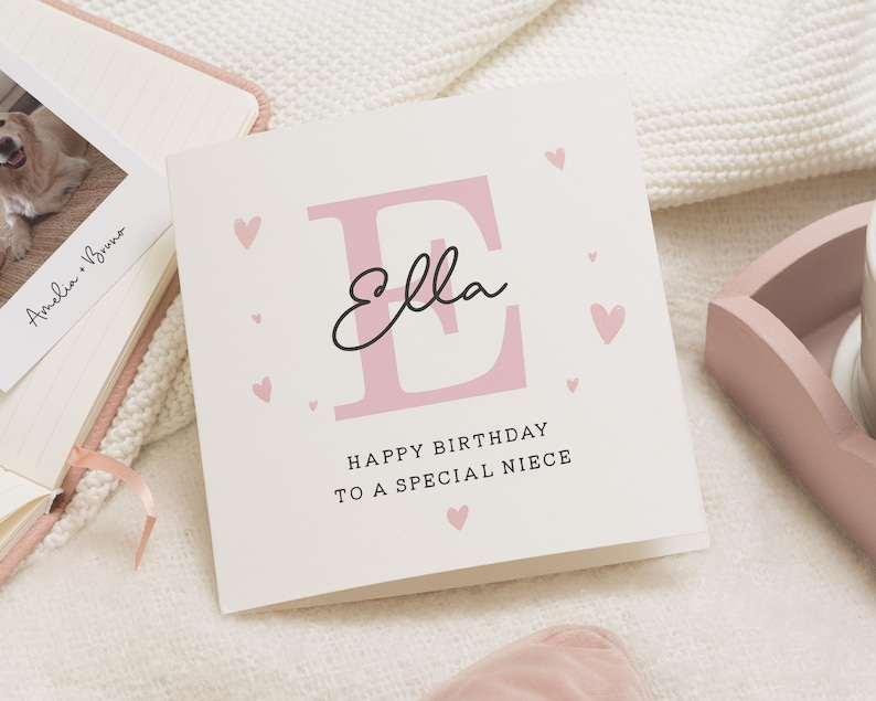 May include: A pink and white birthday card with the letter "E" in pink and the name "Ella" written in cursive. The card says "Happy Birthday to a Special Niece".