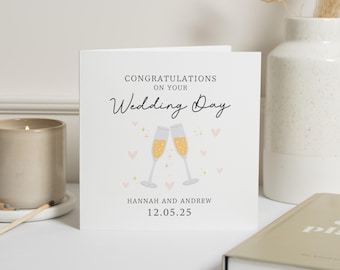 May include: A white card with the text "Congratulations on your Wedding Day" in black lettering. Two champagne flutes with gold liquid are depicted in the center of the card. The names "Hannah and Andrew" and the date "12.05.25" are printed below the flutes.