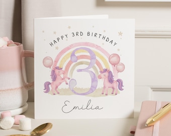 May include: A square birthday card with a watercolor design. The card features a rainbow, two pink unicorns, and the number 3. The text reads "Happy 3rd Birthday" and "Emilia". Two pink balloons are also included.