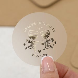 May include: A circular, frosted sticker featuring gold foil details. The text "Grace's Hen Party" is printed around the top edge. Two champagne glasses are illustrated in the centre, with a heart and the date "23.04.26" below.