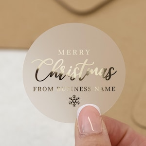 May include: A circular, frosted sticker with gold foil text reading "MERRY Christmas FROM BUSINESS NAME" and a snowflake graphic. The sticker is held by a person's hand.