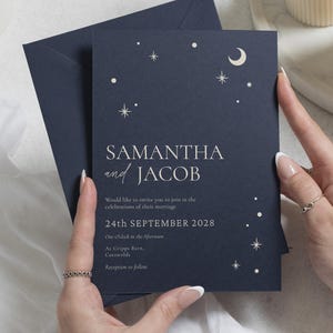 May include: A dark navy blue wedding invitation card and matching envelope featuring white celestial graphics of stars and a crescent moon. The text reads: SAMANTHA and JACOB, Would like to invite you to join in the celebrations of their marriage, 24th SEPTEMBER 2028, One O'clock in the Afternoon, At Cripps Barn, Cotswolds, Reception to follow.
