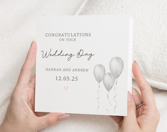 Op de afbeelding: Een witte kaart met de tekst "Gefeliciteerd met jullie huwelijksdag" en de namen "Hannah en Andrew" met de datum "12.05.25". De kaart is versierd met drie zilveren glitterballonnen.