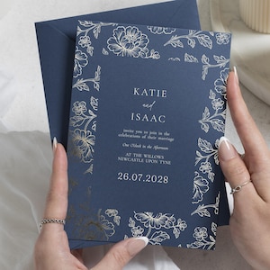 May include: A navy blue wedding invitation card with gold foil floral borders and white text that reads: KATIE and ISAAC invite you to join in the celebrations of their marriage One O'clock in the Afternoon AT THE WILLOWS NEWCASTLE UPON TYNE 26.07.2028.