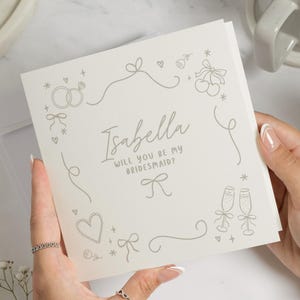May include: An off-white square card with the text "Isabella, Will You Be My Bridesmaid?" in script. The card is decorated with line drawings of rings, bows, hearts, cherries, and champagne glasses. The card is held in two hands.