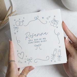 Op de afbeelding: Een witte kaart met de naam "Rosanna" en de tekst "Will you be my something blue?" in blauw schrift. De kaart is versierd met blauwe lijntekeningen van ringen, harten, strikken en champagneglazen. De kaart wordt door twee handen vastgehouden.