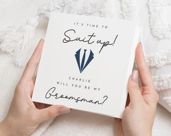 May include: A square, off-white card with the text "IT'S TIME TO Suit up!" in black script. Below, a navy blue tuxedo graphic is centered. The text "CHARLIE WILL YOU BE MY Groomsman?" is written in black script. The card is held by hands.