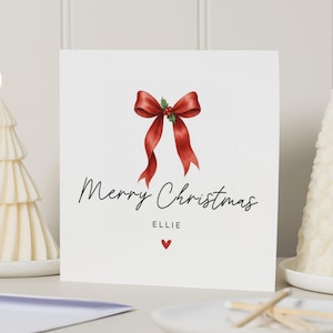Könnte beinhalten: Eine weiße Weihnachtskarte mit einer roten Schleifenillustration und dem Text "Merry Christmas" in schwarzer Schrift. Ein kleines rotes Herz befindet sich unter dem Namen "Ellie". Zwei brennende, weiße, baumförmige Kerzen flankieren die Karte.
