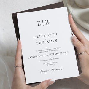 May include: A white wedding invitation with black text, held in front of a dark brown envelope. The invitation reads "ELIZABETH and BENJAMIN" and includes the date "SATURDAY 24th SEPTEMBER 2028". The invitation also includes the text "Reception to follow."