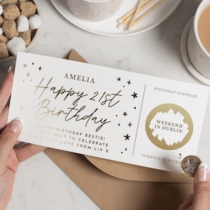 May include: A white rectangular birthday surprise scratch card with gold foil text reading Amelia Happy 21st Birthday and Weekend in Dublin. The card features gold star accents and a perforated line, with a coin held to scratch the gold panel.