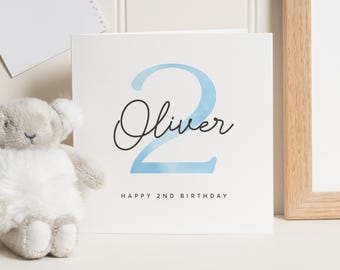 May include: A white card with a blue watercolor number two and the name Oliver written in black cursive. The text "Happy 2nd Birthday" is printed below the name.