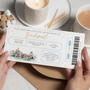 May include: A personalized boarding pass for a trip to Budapest, held by a person. The pass features the text "Pack your bags William, we're going to Budapest" in gold script. It includes passenger details, departure information, and a message.