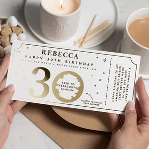 May include: A birthday scratch card with gold foil details. The card says "REBECCA HAPPY 30TH BIRTHDAY" and features a "Trip to Kefalonia" reveal. The card is held over a brown envelope.