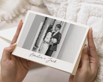 May include: A cream-colored wedding card held by hands, featuring a black and white photograph of a couple kissing. The card is inscribed with the names "Amelia + Jack" in elegant script.