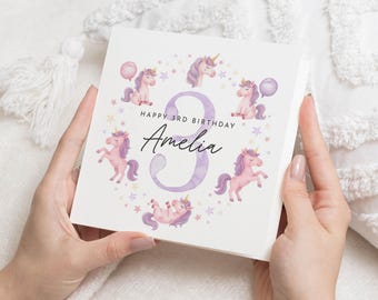 May include: A square birthday card featuring a watercolor design with unicorns, stars, and balloons. The card reads "Happy 3rd Birthday Amelia" with a large number 3. The unicorns are pink and purple.