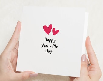 May include: A white greeting card held by hands, featuring two red hearts and the text "Happy You + Me Day" in black. The card is square and has a simple, minimalist design.