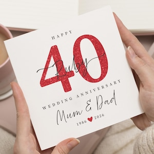 Puede incluir: Una tarjeta blanca con el texto "Happy 40 Ruby Wedding Anniversary Mum & Dad" en escritura negra. El número 40 es en purpurina roja, con la palabra "Ruby" en cursiva. La tarjeta está sostenida por dos manos.