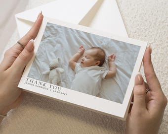 May include: Thank you card with a white background and a photo of a baby sleeping on a white blanket. The baby is wearing a white onesie and has a stuffed animal next to them. The text "THANK YOU" is printed in black at the bottom of the card, along with the baby's name and birthdate.